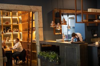 Sala de restaurante com estantes decorativas, mesa com clientes e cozinha aberta com chef a preparar pratos.
