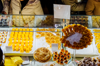 Vitrine com doces amarelos, laranjas e castanhos, com clientes agasalhados do outro lado.