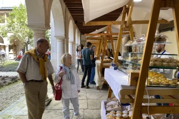 Pessoas caminham por feira coberta em p&aacute;tio com arcos de pedra, observando barracas de p&atilde;es e doces expostos em vitrines sobre mesas brancas.