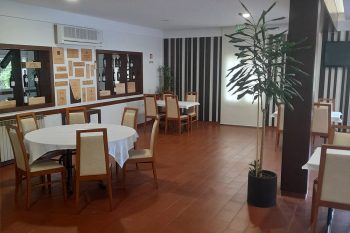 Sala de restaurante com mesas cobertas com toalhas brancas, cadeiras de madeira, expositores de vinho na parede e uma planta decorativa.