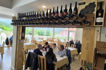 Sala de restaurante com mesas, clientes a comer e garrafeira suspensa com garrafas de vinho.