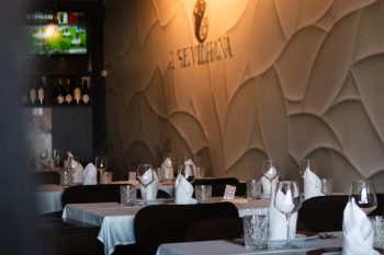 Sala de restaurante com mesas postas, toalhas brancas, copos de vinho e parede decorativa com o nome “A Sevillana”.