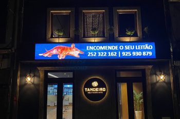 Fachada do Restaurante Tanoeiro à noite, com letreiros iluminados e painel digital com imagem de leitão e contactos.