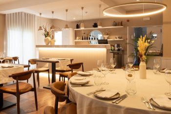 Sala de restaurante com mesas redondas e retangulares cobertas por toalhas brancas, copos, talheres, cadeiras de madeira com assentos em pele, candeeiro circular suspenso e balcão iluminado ao fundo.