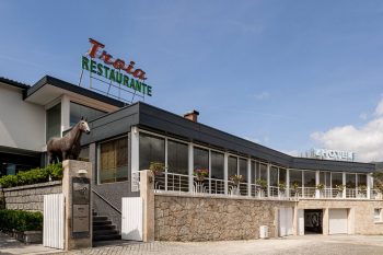 Fachada em pedra e vidro com letreiro “Troia Restaurante” e estátua de cavalo sobre pedestal.