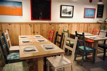 Sala com mesas de madeira, cadeiras pintadas em várias cores, pratos e talheres dispostos; paredes com quadros coloridos e fotografia a preto e branco; iluminação quente e quadro de ardósia com preço.