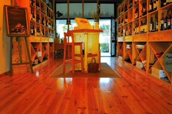 Sala com prateleiras de madeira cheias de garrafas de vinho, mesa central e portas de vidro ao fundo.
