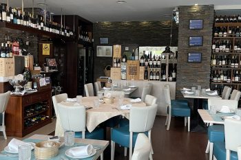 Sala de restaurante com mesas postas, cadeiras brancas e azuis, e garrafeira com várias garrafas de vinho.