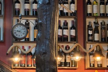 Prateleiras com garrafas de vinho com etiquetas de preço, iluminação quente, relógio antigo e decoração rústica.