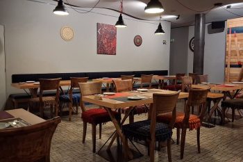 Sala de refeições com mesas de madeira preparadas com pratos, talheres e bases individuais.