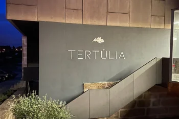 Fachada moderna do restaurante Tertúlia com revestimento metálico, rampa de acesso, jardim com pedras e iluminação noturna. Texto visível: “TERTÚLIA”.