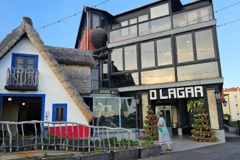 Fachada do restaurante O Lagar com prédio envidraçado de vários andares ao lado de construção com telhado de colmo. Pessoa caminha em frente à entrada decorada com vasos de flores;