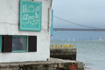Prédio branco com janelas azul-turquesa e placa “Atira-te ao rio Restaurante”, situado na areia da praia com ponte suspensa e veleiros ao fundo.