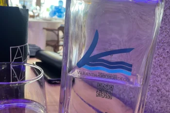 Garrafa de vidro transparente com logotipo "Varanda do Mondego" e QR code sobre mesa de restaurante, ao lado de copo; salão iluminado ao fundo.