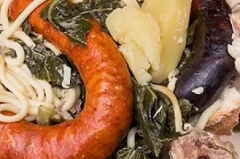 Prato com enchidos, carne cozinhada, massa, legumes verdes e pão.
