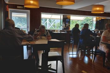 Sala de refeições com várias mesas ocupadas por clientes, iluminação suspensa e janelas amplas.
