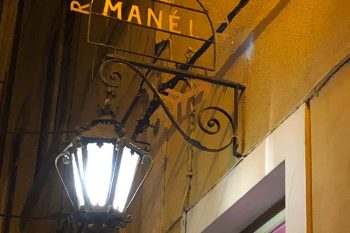 Placa metálica com o nome do restaurante, iluminada por um candeeiro de rua numa parede amarela.